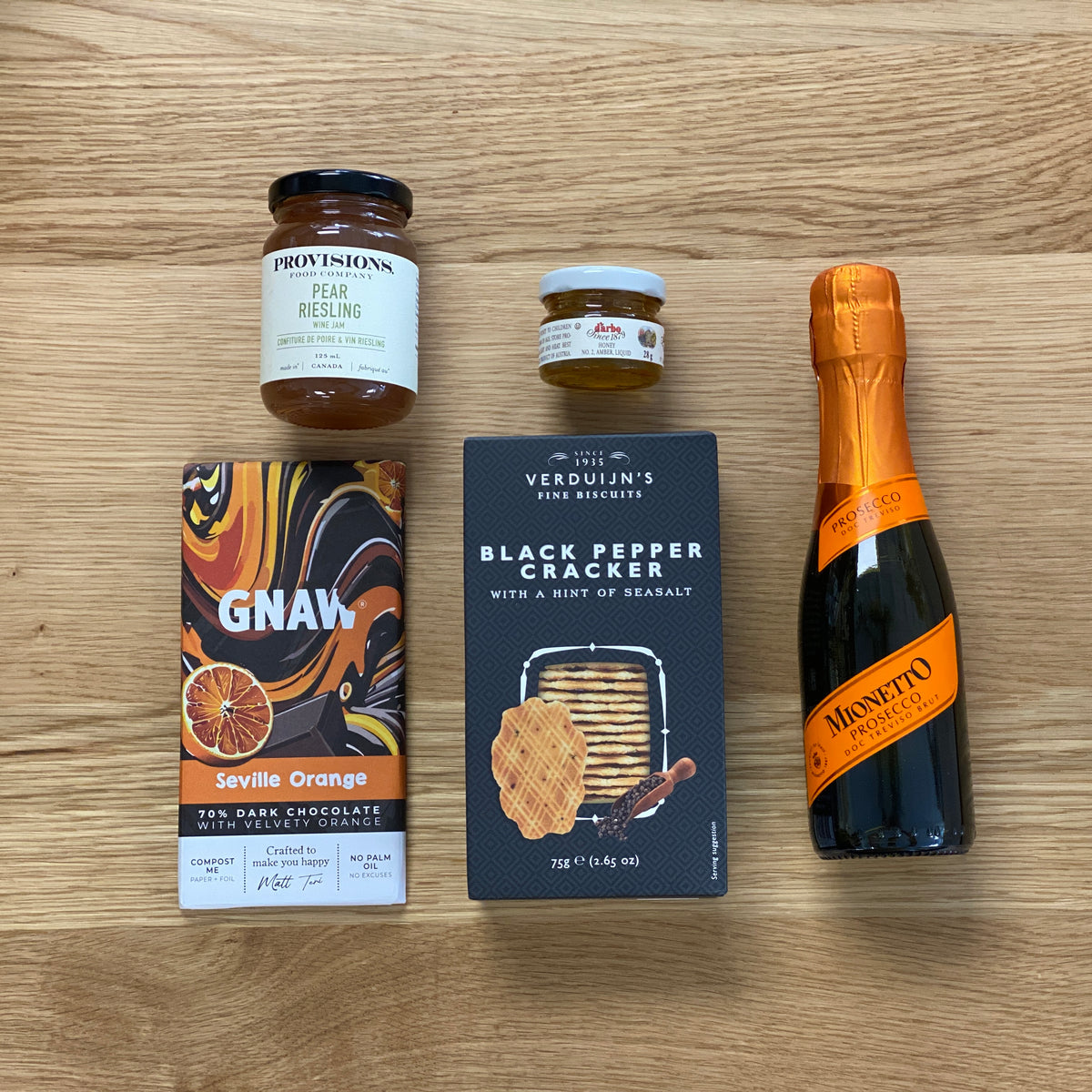 A curated gourmet gift assortment arranged on a light wood tabletop, including a Provisions pear resling jar, a small jar of honey, a GNAW Seville Orange dark chocolate bar, a box of Verduijn's Black Pepper Cracker, and a Mionetto sparkling wine bottle.