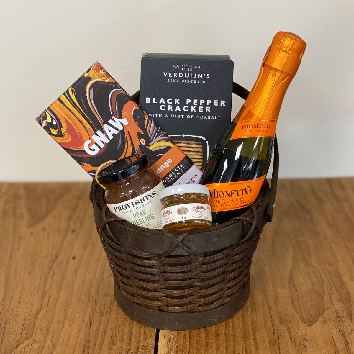 A gourmet gift basket in a dark brown wicker basket, filled with assorted snacks and treats including a bottle of Prosecco, a box of black pepper crackers, a bag of GNAW chocolate, and jars of Provisions Pear Reisling and a Honey jar, set on a wooden table against a neutral background.